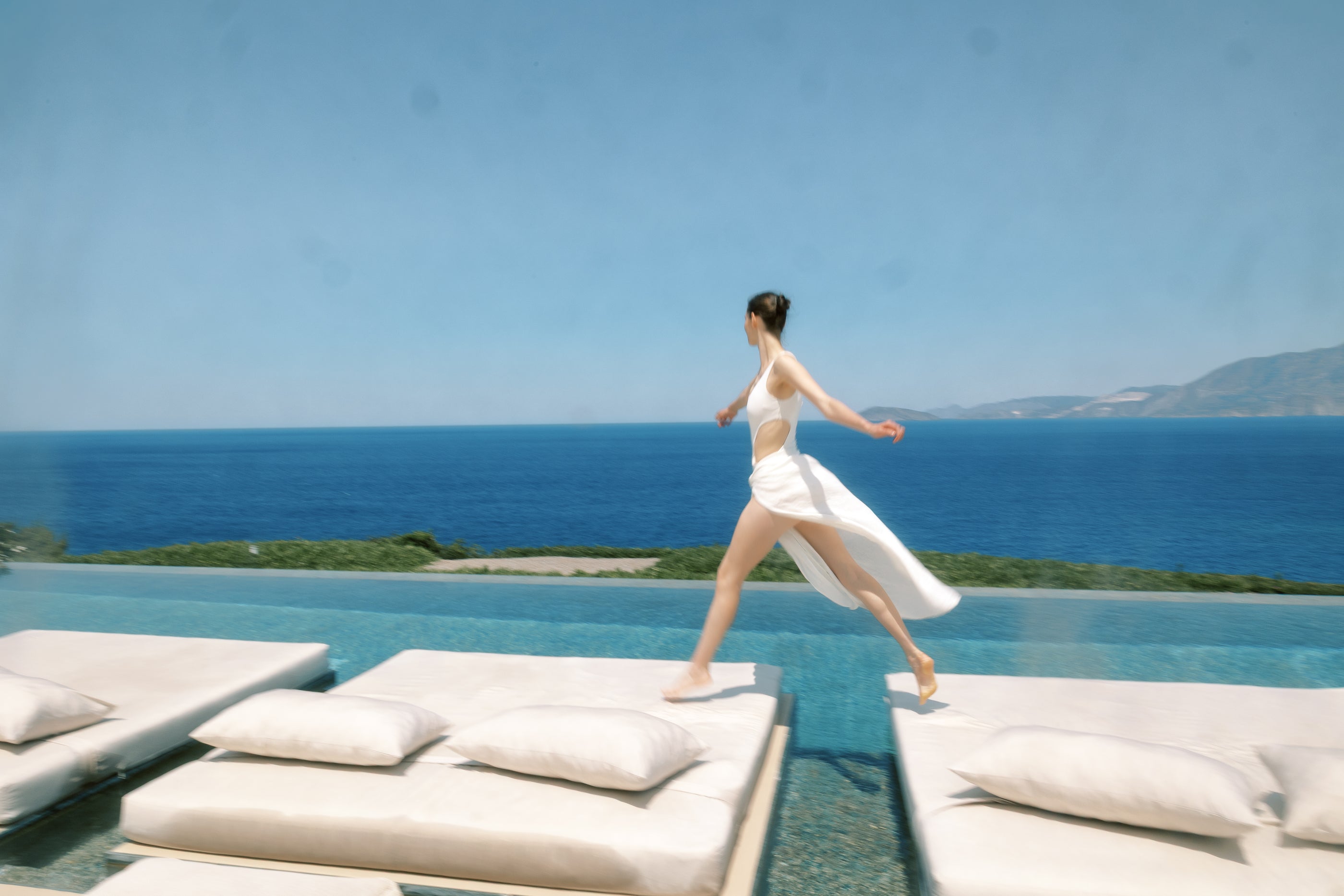 Woman in a white dress standing on a sunbed by a pool with a scenic ocean view.