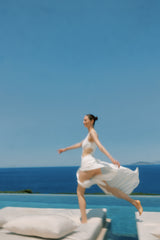 Woman in a white dress standing on a pool edge with a clear blue sky and ocean background