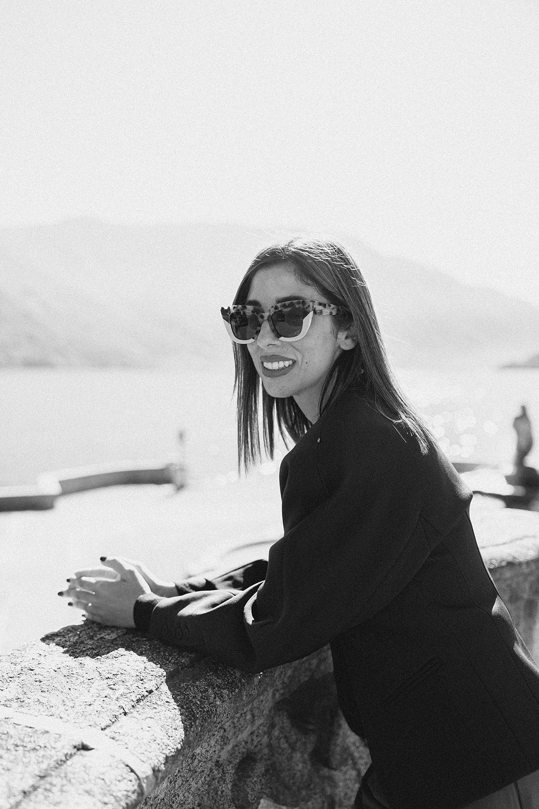 Black and white photo of a woman wearing sunglasses and a dark outfit, standing on a rocky surface with water in the background.