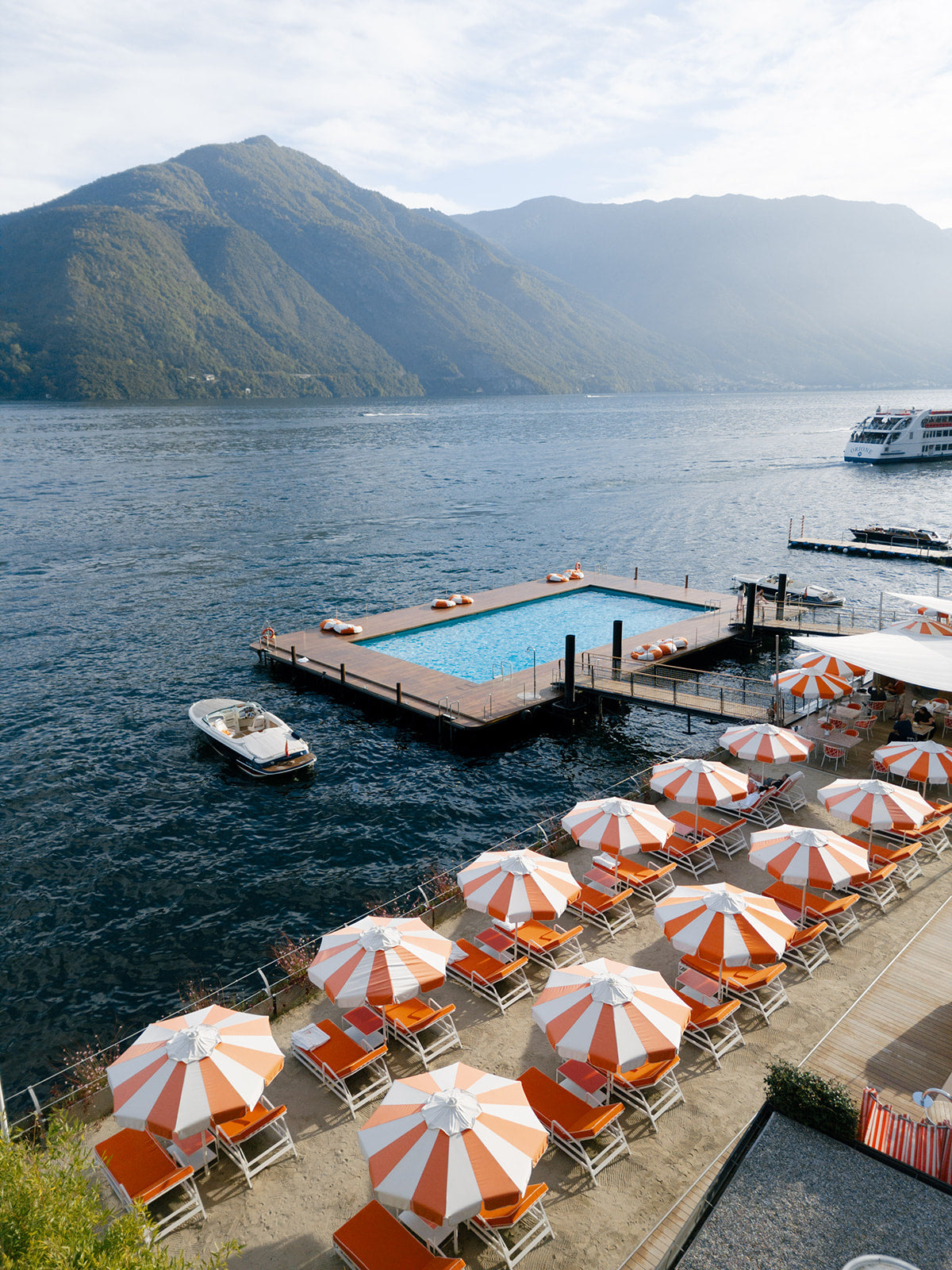 Outdoor seating area with orange and white umbrellas by a lake with mountains in the background Grand Hotel Tremezzo