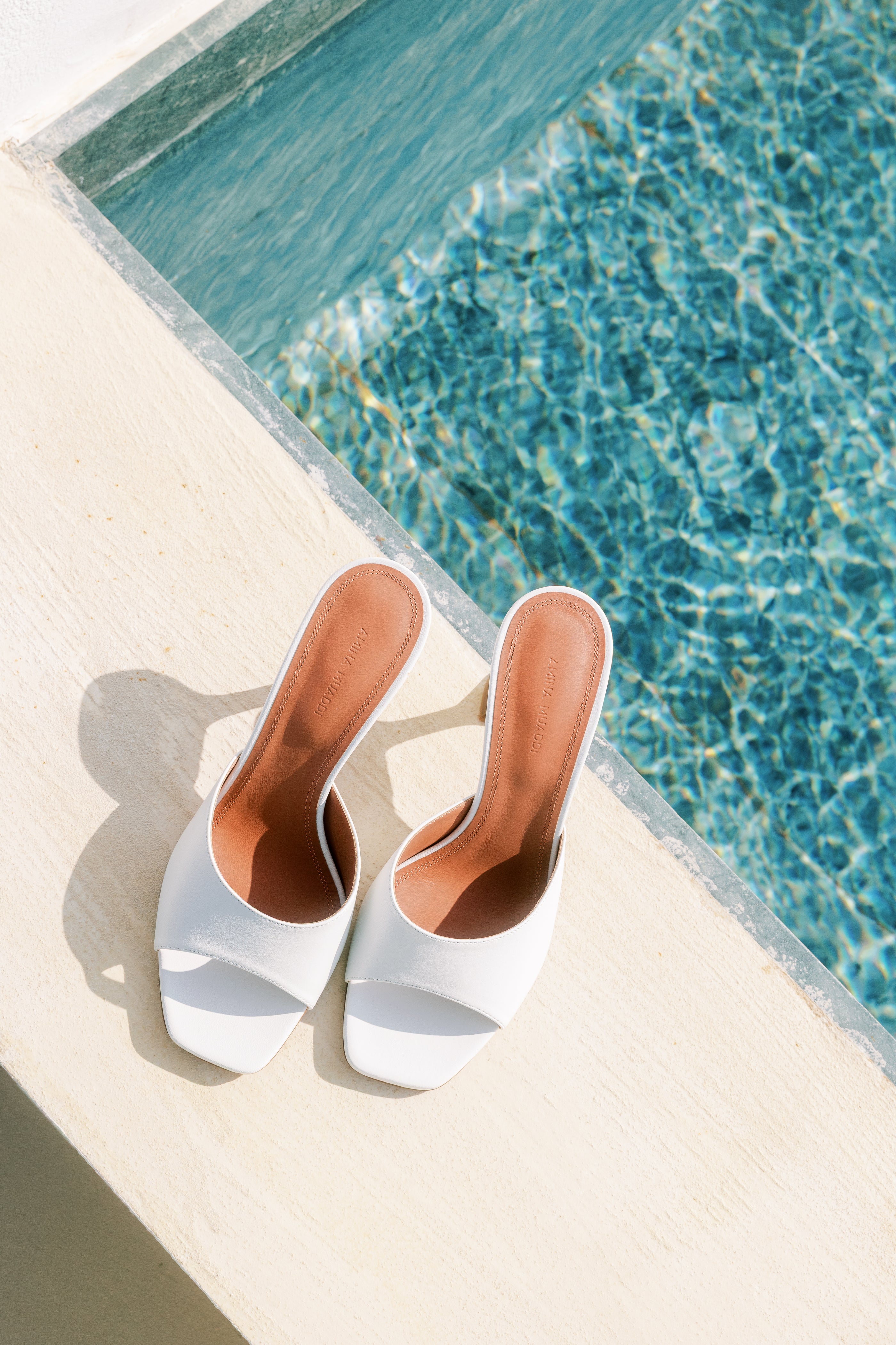 White high-heeled shoes Amina Muaddi with brown soles on a poolside ledge.